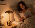 A woman reading a book in bed under a warm bedside lamp, with a gold-toned table lamp on the nightstand beside her.
