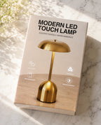 A boxed modern LED touch lamp in a warm metallic gold finish, photographed on a light marble surface with soft sunlight and shadows. The box reads 'MODERN LED TOUCH LAMP' and shows a matching lamp illustration on the front.